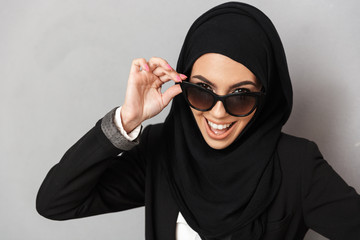Portrait closeup of stylish muslim woman 20s in religious headscarf smiling and holding sunglasses, isolated over gray background