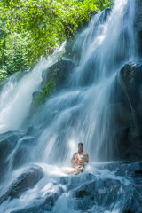 attractive woman sitting at rock in yoga pose for spiritual relaxation serenity and meditation at stunning beautiful waterfall and rain forest in Bali