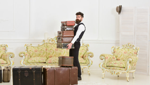 Macho elegant on strict face carries pile of vintage suitcases. Butler and service concept. Man with beard and mustache wearing classic suit delivers luggage, luxury white interior background.