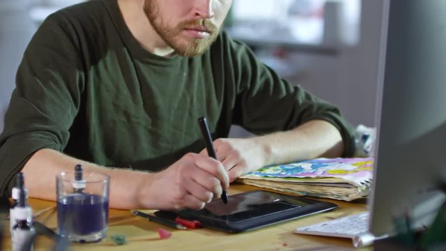 Young male designer using stylus and drawing tablet connected to computer to create illustrations