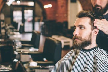 Bearded guy is sitting in salon. His hairstylist is modelling his hair. Guy is looking straight forward.