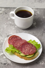 cup of black coffee and sandwich with sausage, cucumber and salad