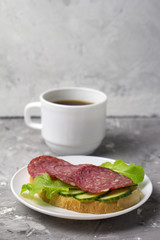 cup of black coffee and sandwich with sausage, cucumber and salad