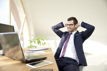 Financial consultant businessman. Portrait of young business man in suit and glasses using laptop while sitting with hands on his head and working on new project. 