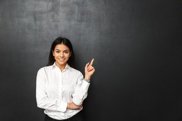 Portrait of secretary or business woman with long dark hair wearing white shirt pointing finger upward on copy space, isolated over dark gray background