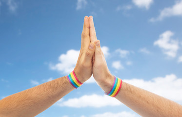lgbt, same-sex love and homosexual relationships concept - close up of male couple hands with gay pride rainbow awareness wristbands making high five gesture over blue sky and clouds background