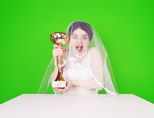 Caucasian bride in a wedding attire holding a golden trophy. Half length of young groom with a trophy. Happy bride in fot sitting at a table celebrating with a trophy on a green background.