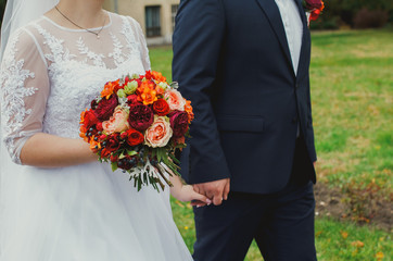 Plus size wedding couple is walking outside. Curvy bride is holding beautiful colorful bouquet with orange, red and pink peonies and roses. Bride and groom in summer green love story.