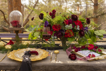 A red rosebud lying on a gold platter with a black veil, next to a glass of red wine, golden spoon and knife, golden fruits and flowers on an old wooden table