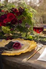 A red rosebud, lying on a golden platter, next to a glass of red wine, a golden spoon and a knife, golden fruits and flowers on an old wooden table