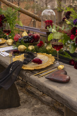 A red gold bud lying on a golden plate with a black veil, next to a glass of red wine, a wooden heart, golden fruits and flowers on an old wooden table.