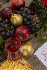 A red rosebud, lying on a golden platter, next to a glass of red wine, a golden spoon and a knife, golden fruits and flowers on an old wooden table