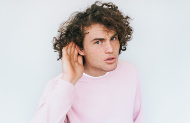 Handsome interested male with curly hair placing hand on ear asking someone to speak up, isolated...