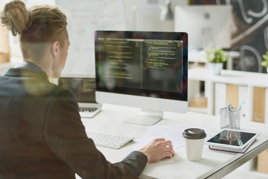 Rear view of hipster coder with hair bun wearing jacket using computer while programming app and sitting at desk with technologies and coffee cup