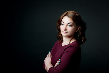 A brown-haired girl with a make-up and hairdo in a claret dress crossed her arms over her chest against a black background.