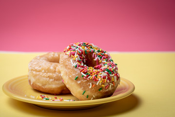 Two glazed doughnuts with colorful sprinkles on a yellow plate with pink background.