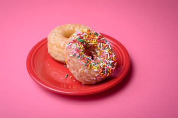 Two glazed donuts, one with colorful sprinkles, on a red plate with a pink background.