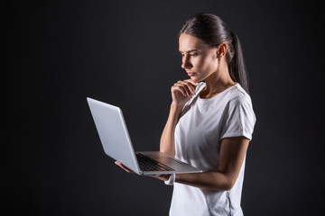 Focused on thoughts. Smart pleasant attractive woman holding a laptop and looking at its screen while touching her chin