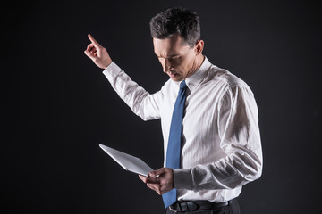 Personal gadget. Pleasant nice adult man standing and pointing at the sensory panel while looking at the tablet screen