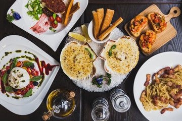Italian dishes on the table including Burrata Salad, Baked Cod with sauce,  Spaghetti Carbonara, Baked scallops with cheese and Canapes with olive oil, pepper and salt on the table.