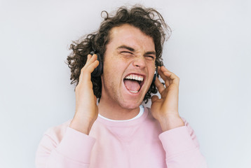 Closep shot of freedom handsome stylish young male with curly hair wearing pink clothes, singing and listening favorite music on headphones, isolated on white studio wall. Copy space for advertisement