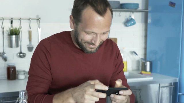 Happy, young man playing game on smartphone sitting by table at kitchen
