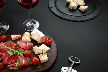 Delicious appetizer to wine - ham, cheese, baguette slices, tomatoes, served on a wooden board, and glass with red wine on black surface