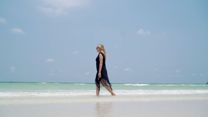 Fototapeta na wymiar A beautiful girl in a black tunic is standing in the water in the ocean. The girl is walking along the ocean.