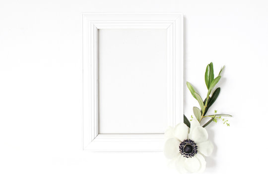 White blank wooden picture frame mockup with green olive branch and anemone flower lying on the white table. Poster product design. Styled stock feminine photography. Home decor. Spring concept.