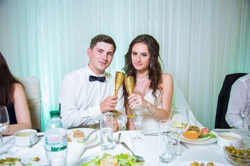 Bride and groom clink glasses of gold with champagne, sitting on the wedding table