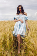 woman in a blue light dress stands in a field with yellow dry ears of wheat in the summer