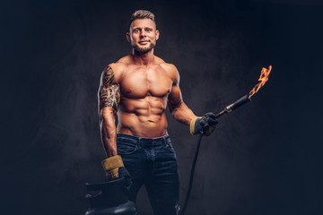 Fototapeta na wymiar Brutal tattoed male welder with a stylish haircut and beard, with muscular body, dressed in only jeans, holds propane tank and a burning burner, standing in a studio, looking at a camera.