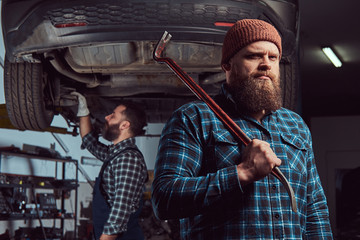 Two bearded brutal mechanics repair a car on a lift in the garage. 