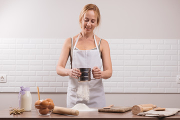 Young woman sifting flour through a sieve on  wooden table. The chef in white apron sifts the flour through a sieve to prepare the dough for pizza on a light background.