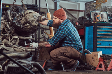 A brutal tattooed bearded mechanic specialist repairs the car engine which is raised on the hydraulic lift in the garage. Service station.