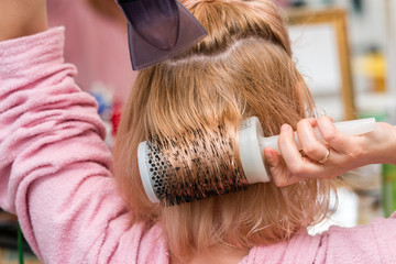 Fototapeta premium Woman drying her long blond hair with hair dryer and round brush. Close-up.