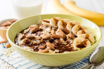 Oatmeal porridge with banana, almonds and chocolate syrup on white wooden background