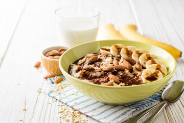 Oatmeal porridge with banana, almonds and chocolate syrup on white wooden background