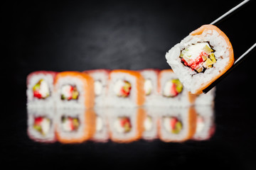 Chopsticks holding sushi roll Philadelphia on black background made of salmon, tuna, cucumber, nori, pickled rice, masago caviar, avocado, lemon. Sushi menu. Japanese food. Free-space.
