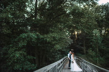 Handsome groom in a tuxedo and a beautiful bride in a white wedding dress are standing on an old wooden bridge in the forest. Bride holds a wedding bouquet from different flowers