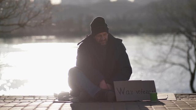 Homeless disabled war veteran male sitting on the street with cardboard sign and paper cup, begging for money. Bearded mature beggar staring at people walking and passing by near the river