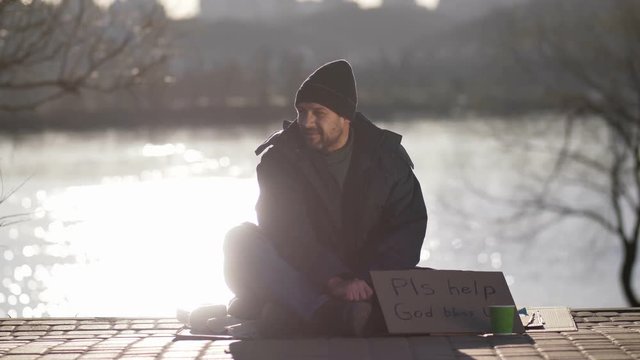 Mature beggar man sitting on pavement at river bank with cardboard sign asking for help. Bearded homeless man in hat looking at people passing by. Woman stops and throws some change in paper cup