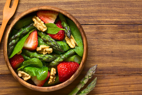 Fresh strawberry, green asparagus, baby spinach and walnut salad served in wooden bowl, photographed overhead on dark wood with natural light (Selective Focus, Focus on the top of the salad)