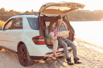 Beautiful young couple with map sitting in car trunk near river