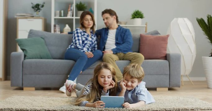 Two pretty cute kids, son and daughter, lying on the carpet in the living room and watching something funny on the tablet device. Their parents sitting on the couch behind them. Indoors