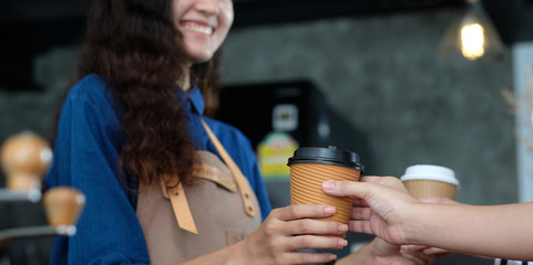 Young asia woman barista holding a disposable coffee cup with smiling face at cafe counter background, small business owner, food and drink industry