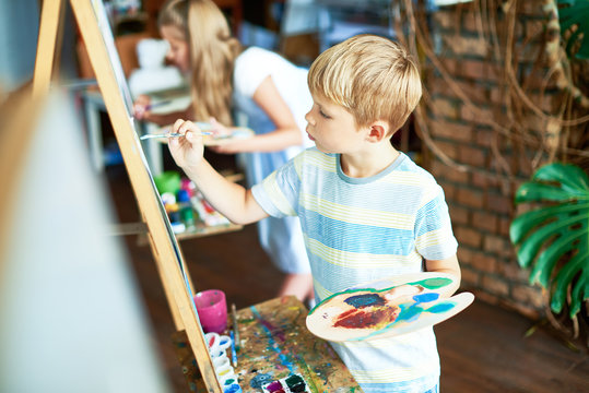 Side view  portrait of blonde little boy painting on easel enjoying art class with other children in background