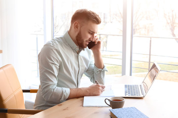 Young man talking on mobile phone while working with laptop at desk. Home office