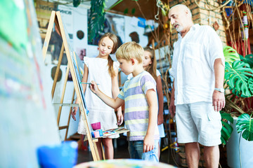 Portrait of senior teacher watching  little kids painting picture in art studio standing by easel with children