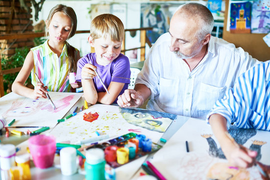 Group portrait of three happy children painting pictures at table in art class with senior teacher
